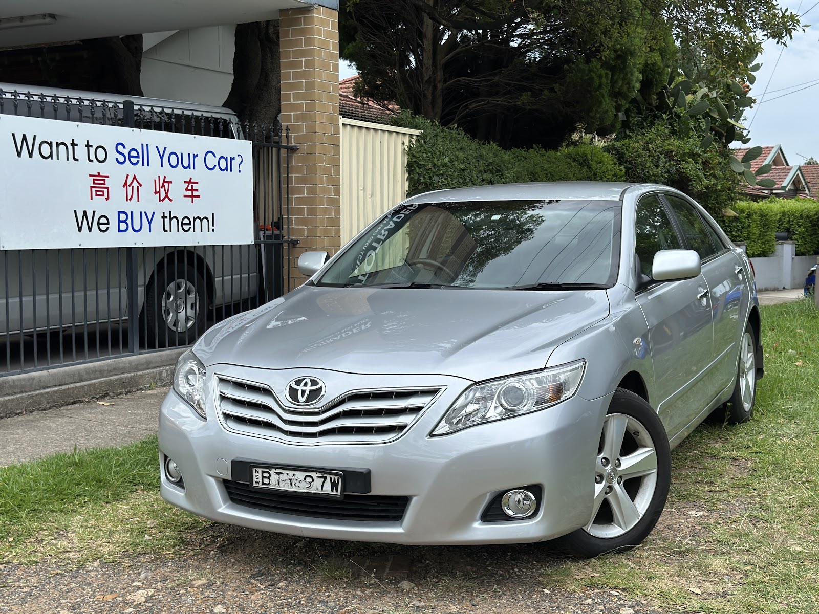 A sign at the yard reading Want to Sell Your Car? 高价收车 We BUY them! beside a silver Toyota Camry